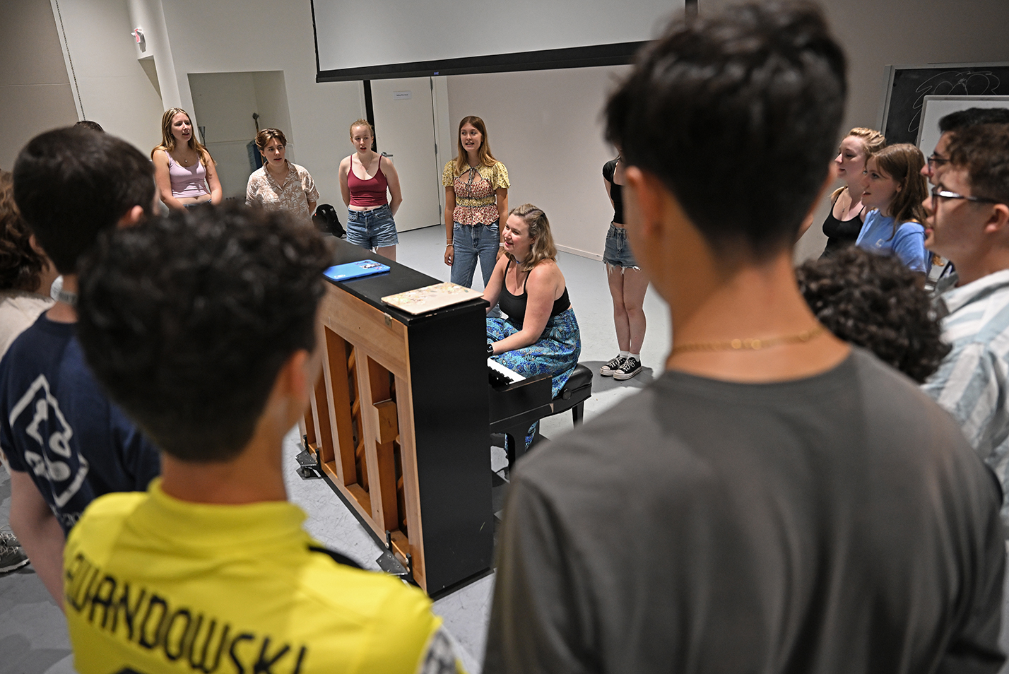 Students gather around a piano to sing.