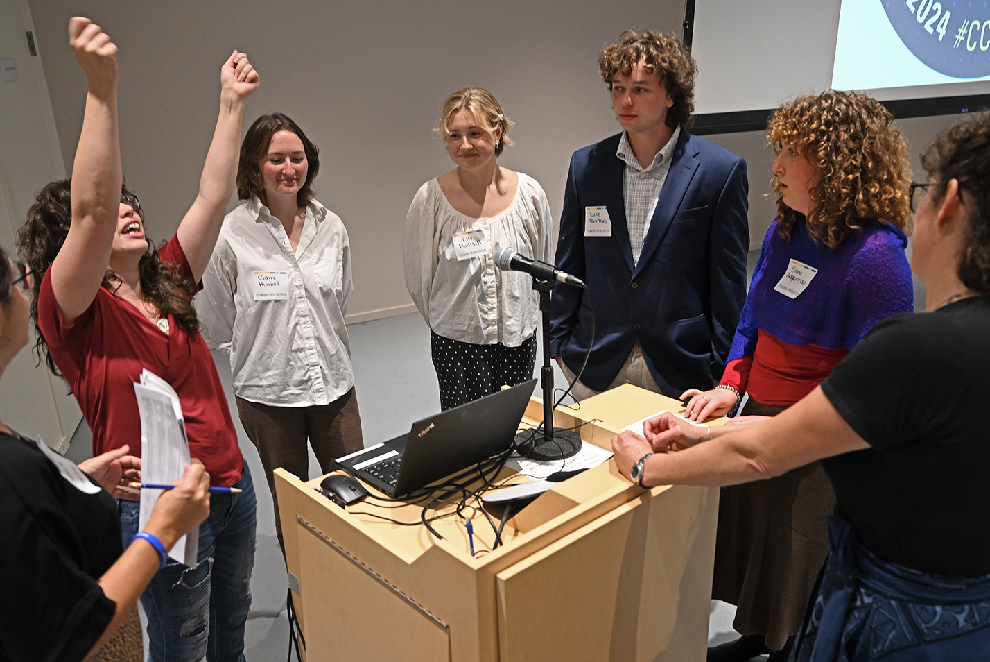 a staffer explains the operation of presentation technology to a group of student speakers around a podium.