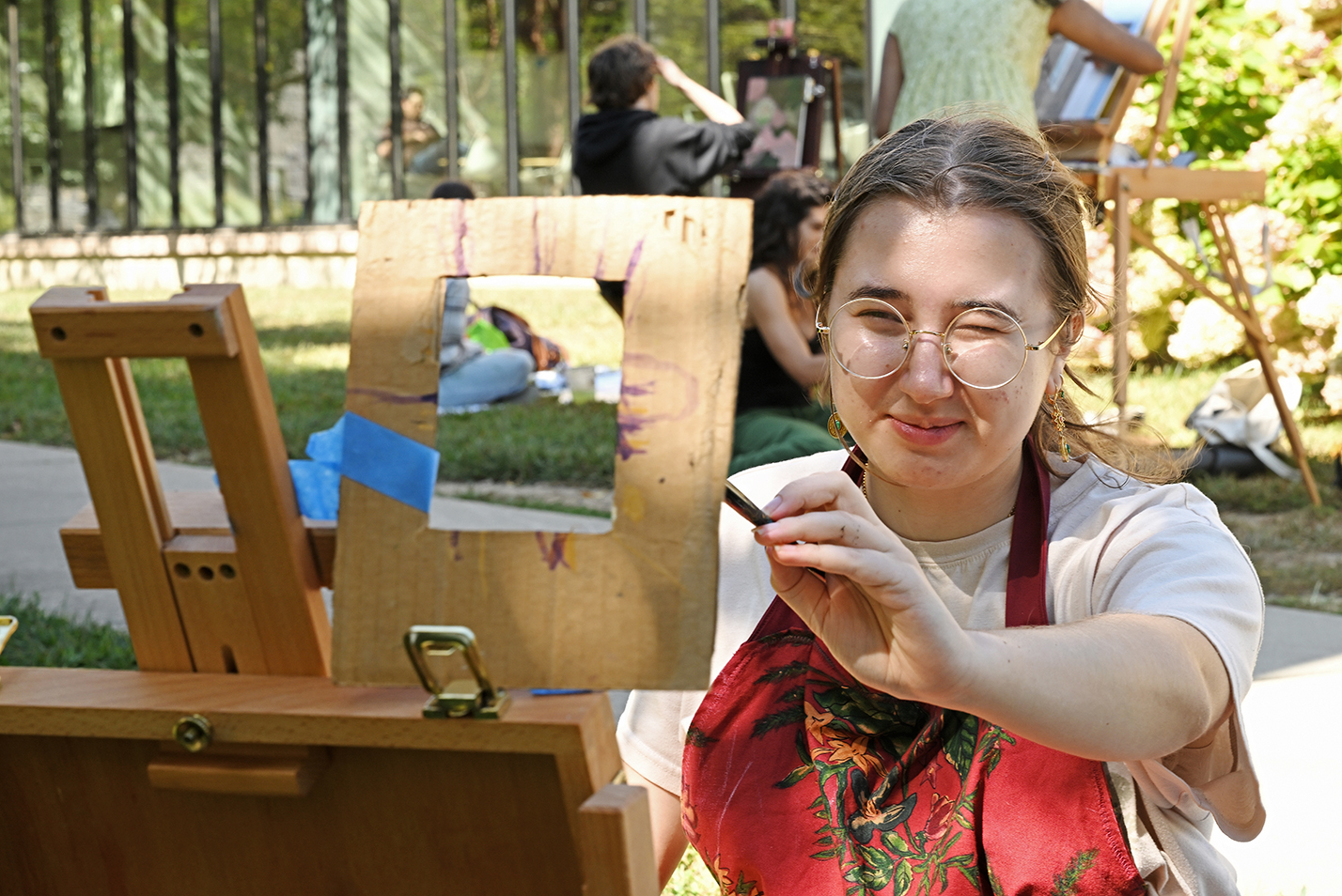 A student uses a frame to compose as she paints on an easel outdoors.