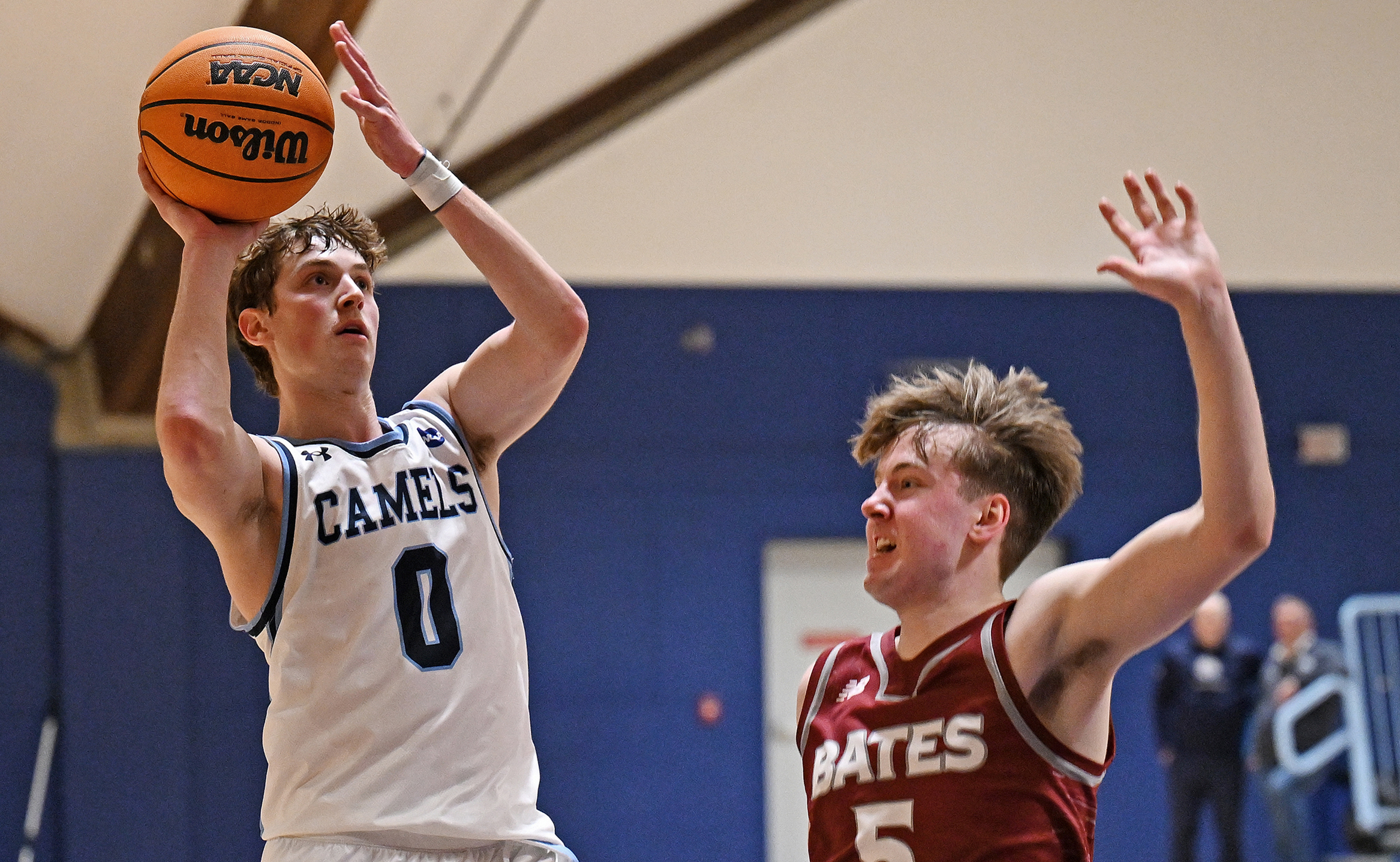 A basketball player in white and blue shoots the ball over a defender in red and white.